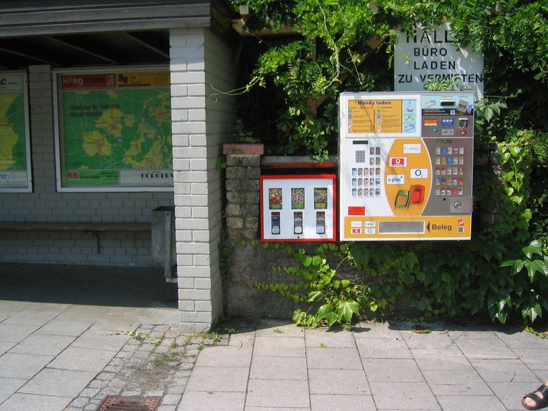 Rot-weißer Automat an einer Gartenmauer neben einem Schaukasten