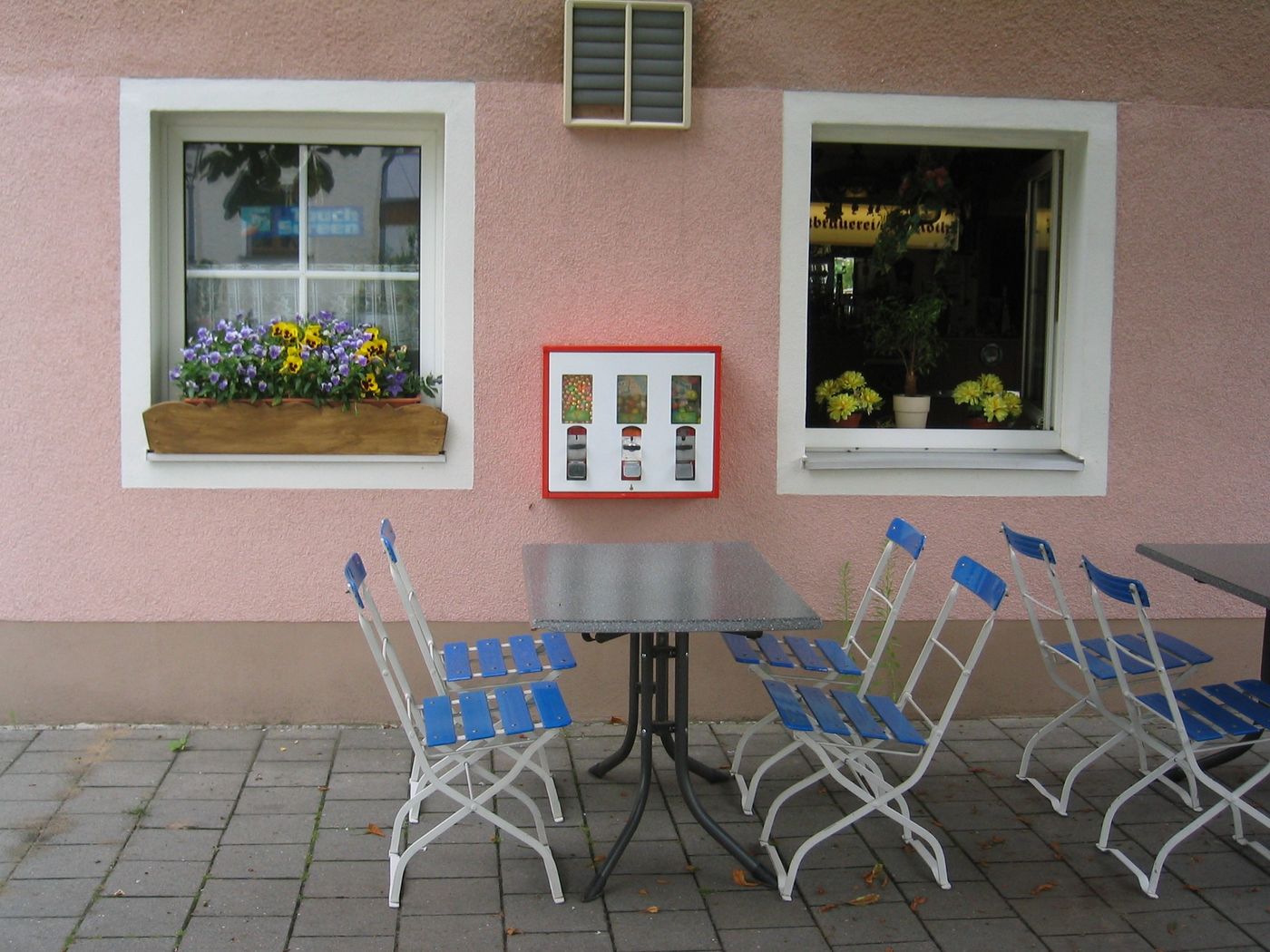 Rot-weißer Automat an einer rosa Hauswand über einem Biertisch mit Klappstühlen