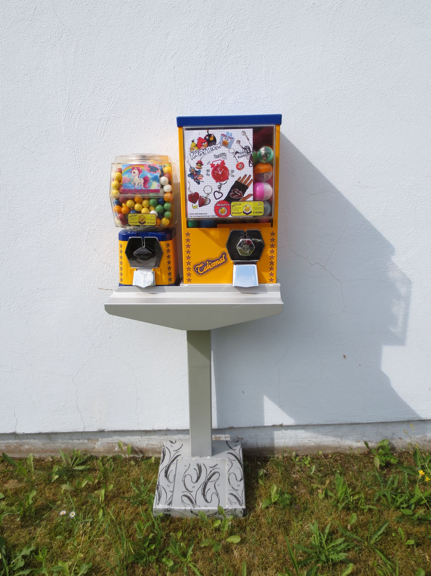 Talismat und Kaugummiautomat auf einem Standfuß vor weißer Wand